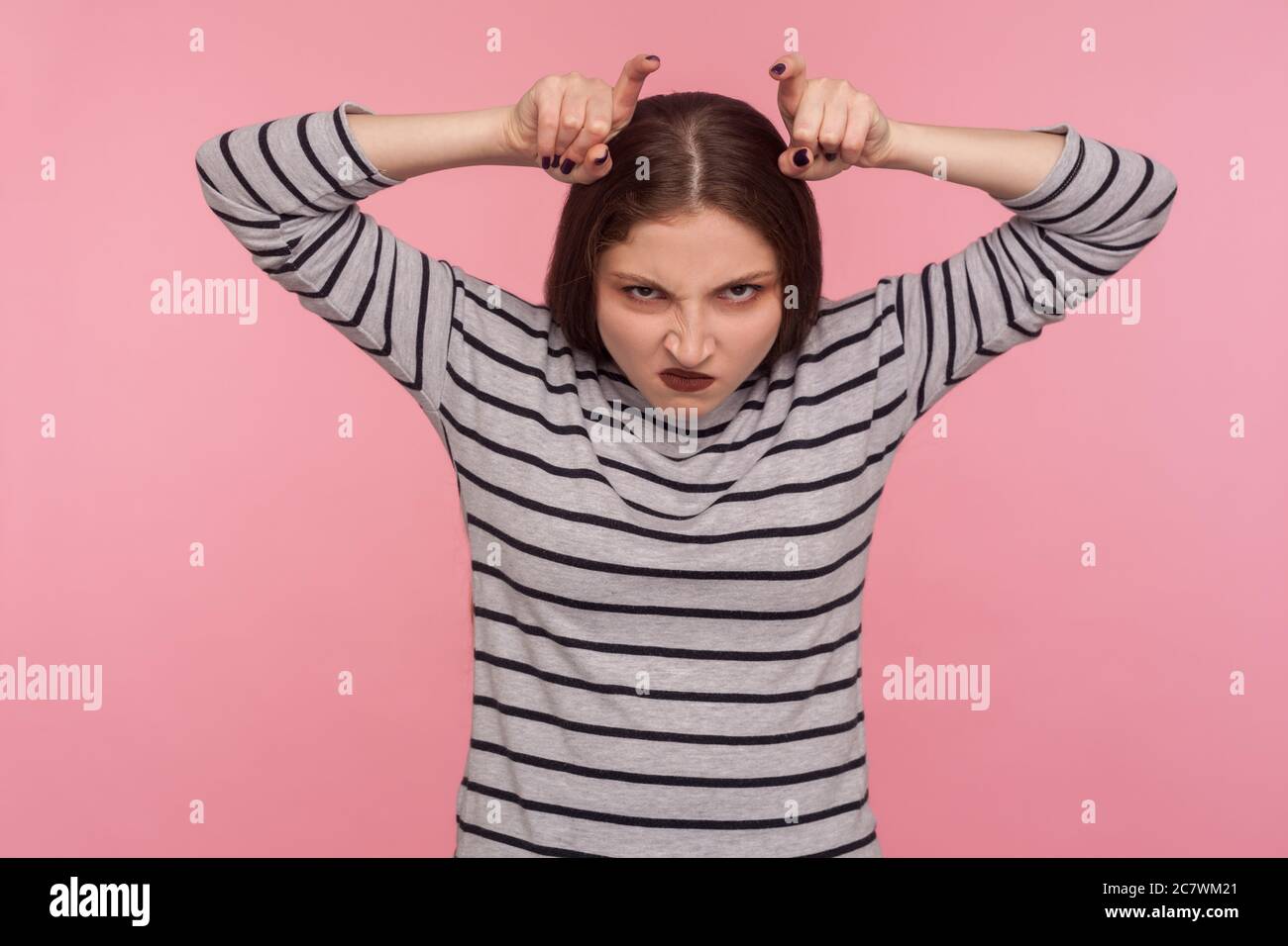 Portrait d'une femme en colère drôle dans un sweat-shirt rayé montrant le mouvement de corne de taureau et regardant avec la rage de haine à la caméra, menaçant et provoquant des conflits Banque D'Images