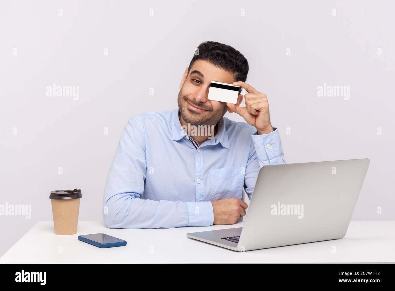Un joueur positif assis sur le lieu de travail avec un ordinateur portable, couvrant l'œil avec une carte de crédit et souriant. E-commerce et services bancaires, concep d'achat en ligne Banque D'Images