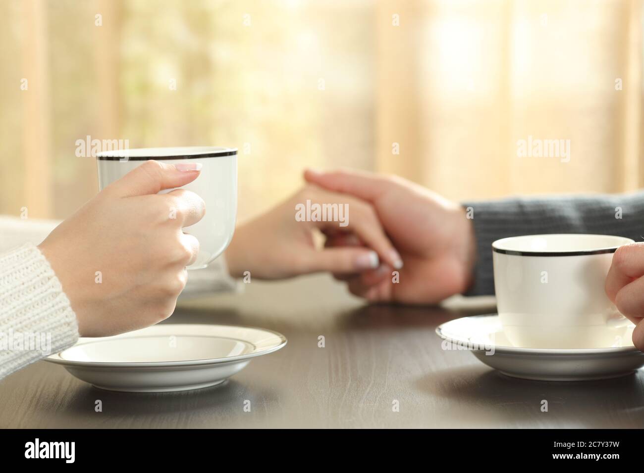 Gros plan d'un couple tenant les mains avec des tasses de café assis sur une table à côté de la fenêtre à la maison Banque D'Images
