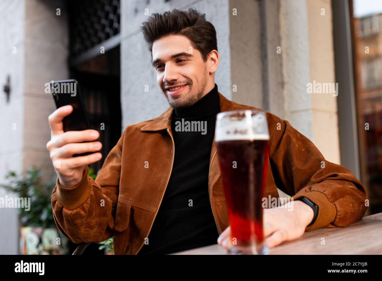 Jeune homme décontracté attrayant utilisant heureusement le téléphone portable tout en buvant de la bière dans un café dans la rue Banque D'Images