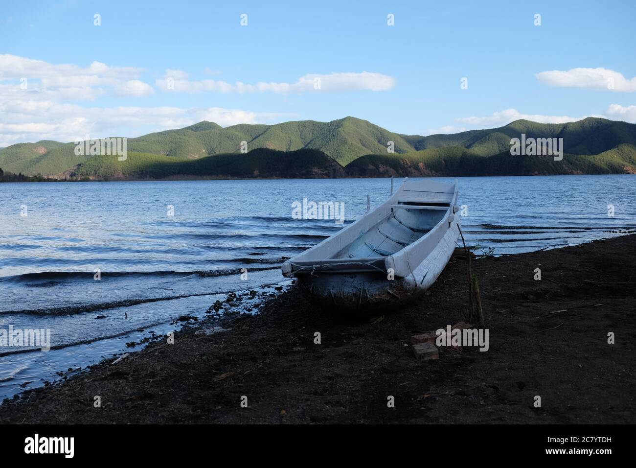 une rangée de bateaux blancs sur la plage de sol noir. Le lac bleu se déforme. Fond vert des montagnes. Ciel bleu ensoleillé. En Lugu lac Sichuan Chine. Banque D'Images