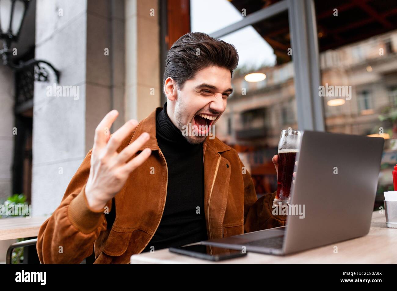 Jeune homme émotionnel positif hurlant joyeusement en travaillant sur ordinateur portable dans un café dans la rue Banque D'Images