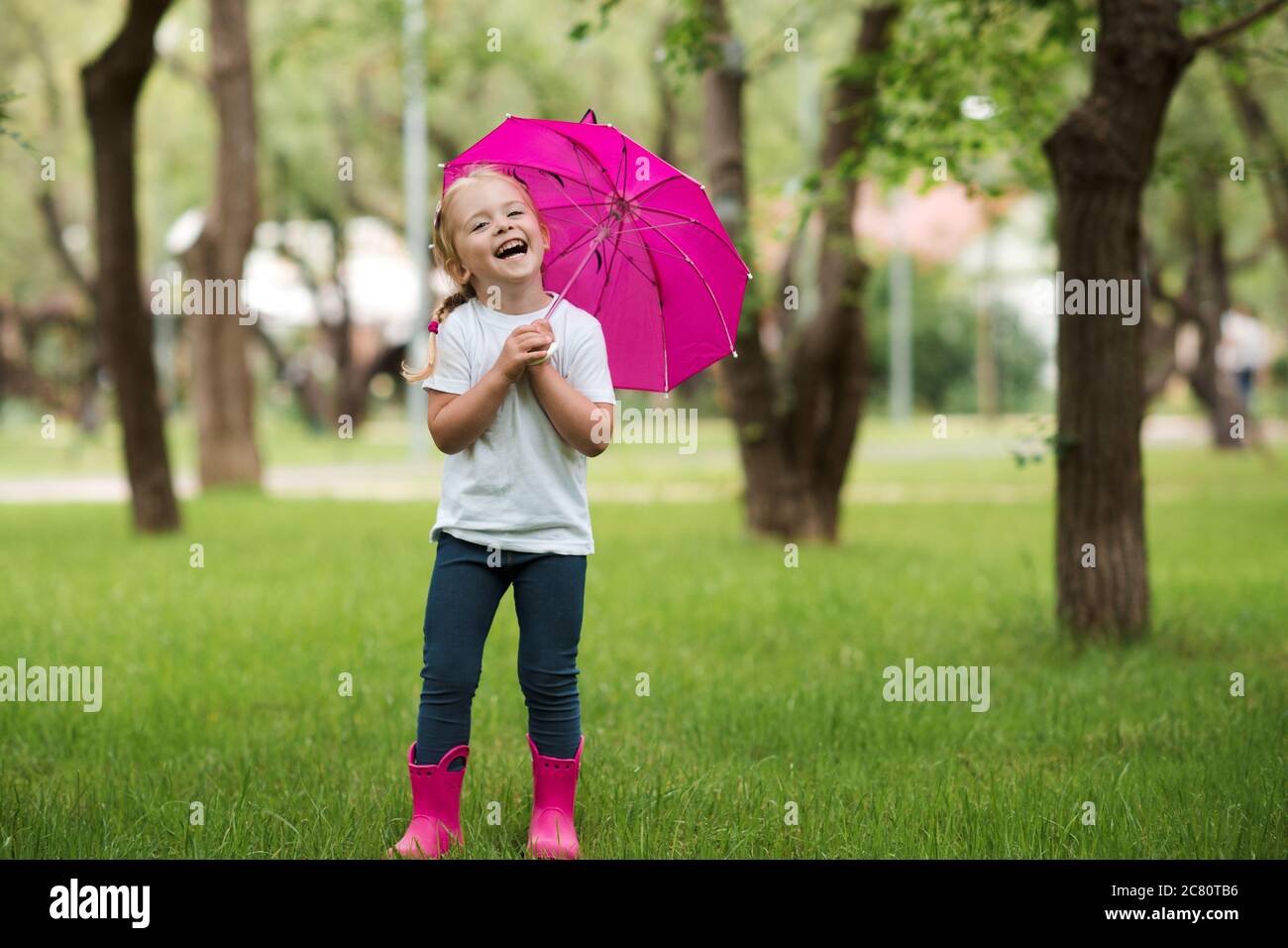 Bonne rigolante fille de 2-3 ans portant des bottes imperméables et tenant  un parapluie rose posant dans le parc sur l'herbe verte à l'arrière-plan.  Bonheur. Loo Photo Stock - Alamy