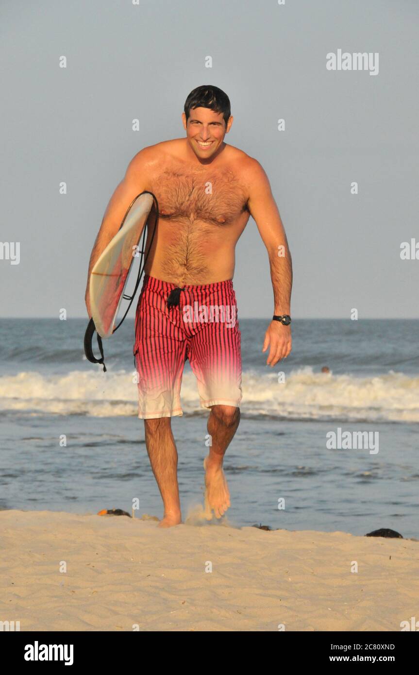 Portrait d'un jeune homme beau dans ses années vingt à venir hors de l'océan transportant une planche de surf sous son bras et souriant Banque D'Images