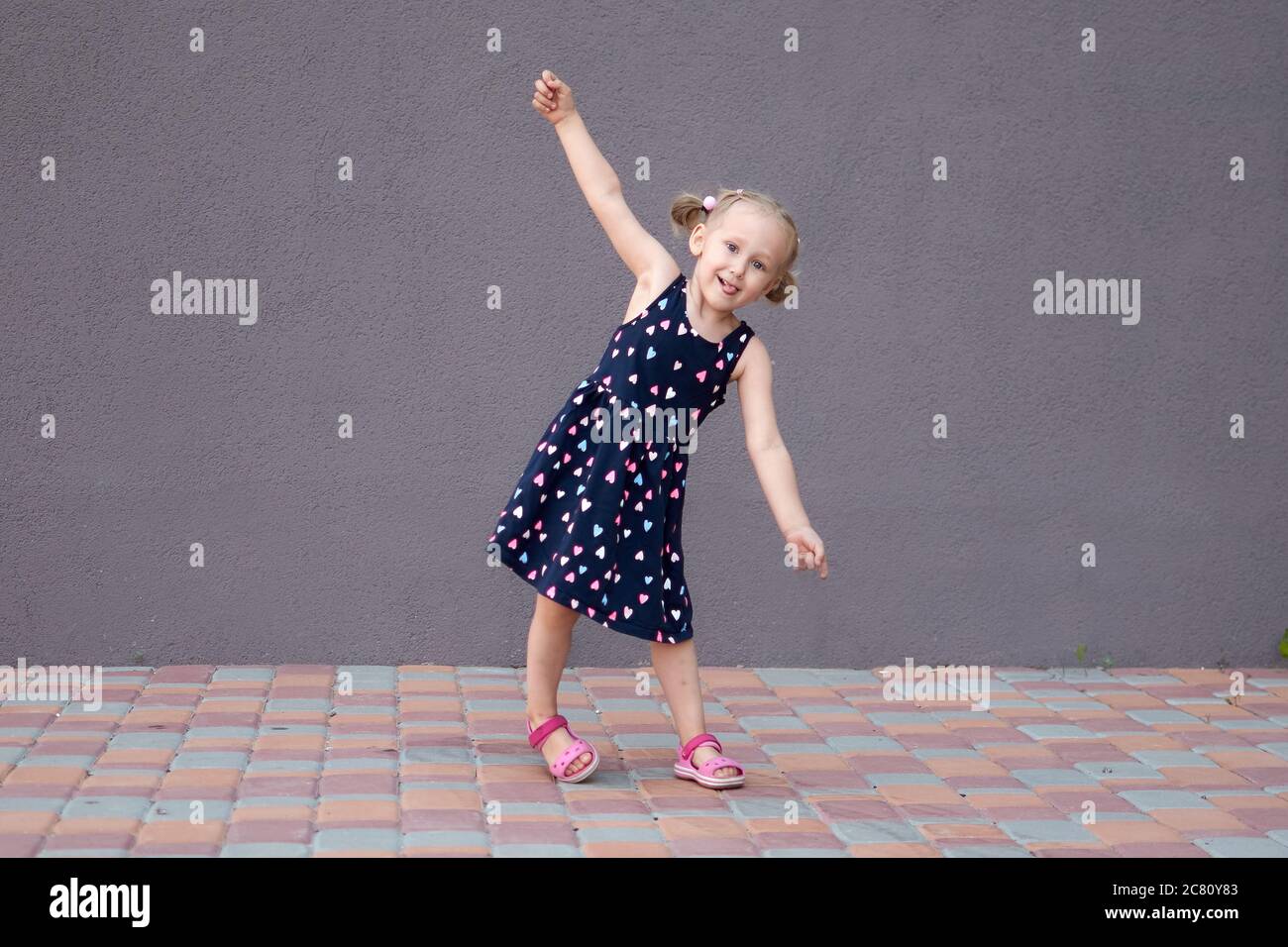 Portrait d'une heureuse liitle blonde fille gros plan. 3 yaers une fille de crapaud caucasienne souriant à la marche à l'extérieur. Arrière-plan de mur en pierre avec espace de copie. Enfant Banque D'Images