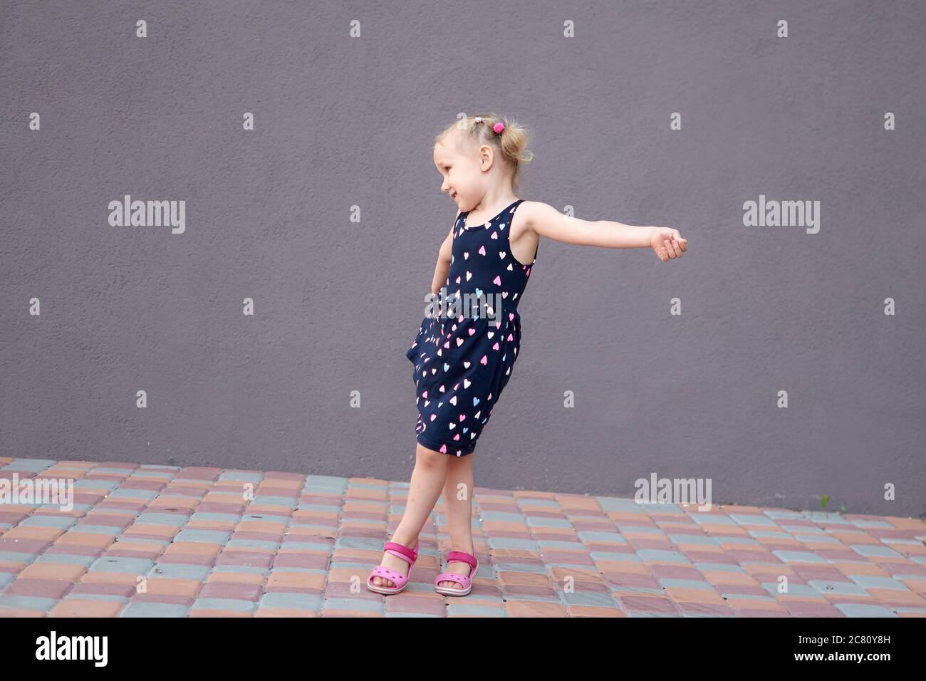 Portrait d'une heureuse liitle blonde fille gros plan. 3 yaers une fille de crapaud caucasienne souriant à la marche à l'extérieur. Arrière-plan de mur en pierre avec espace de copie. Enfant Banque D'Images