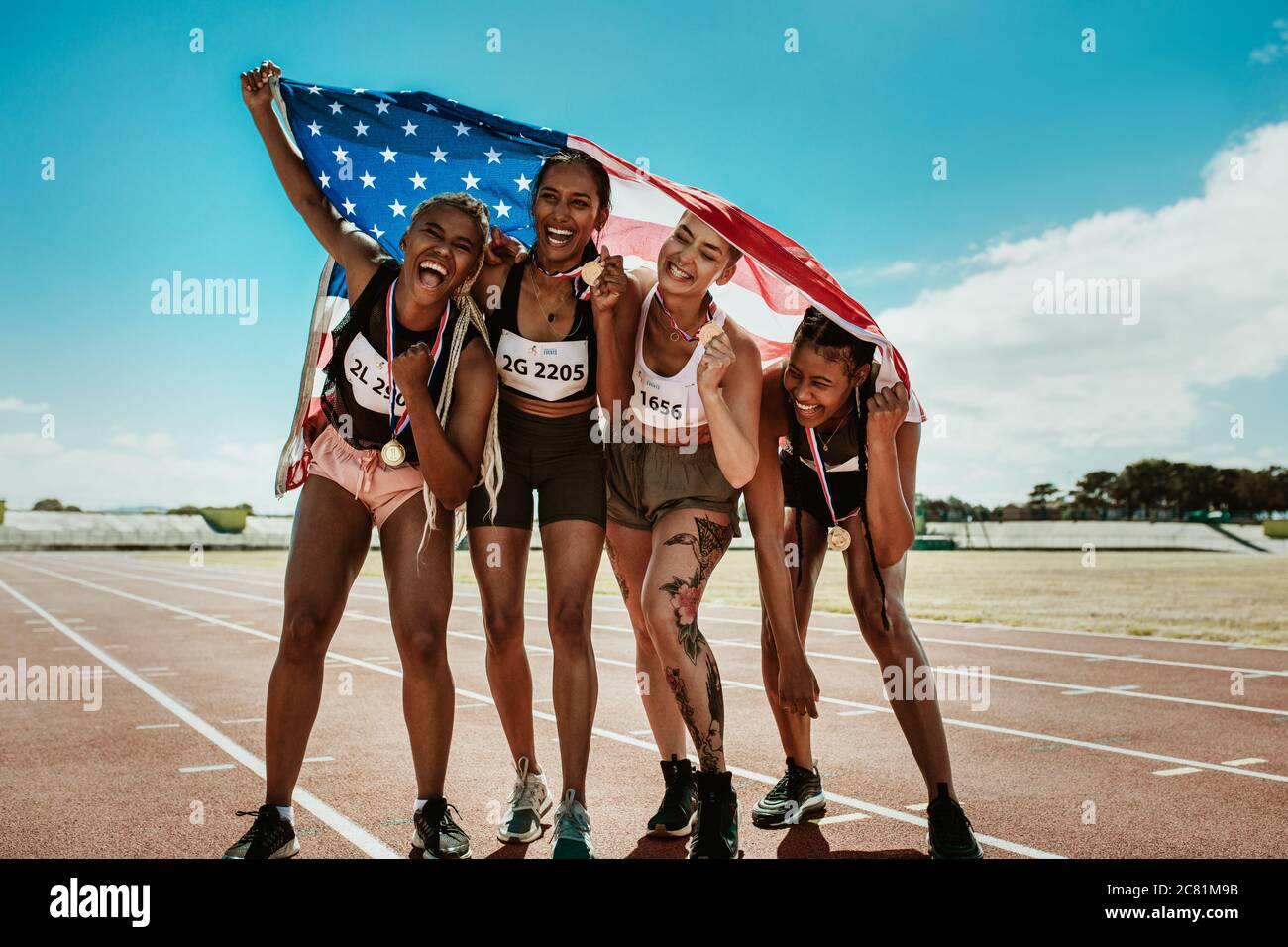 Portrait de la jeune équipe d'athlètes féminins profitant de la victoire. Groupe de coureurs divers avec des médailles célébrant le succès en tenant un drapeau des États-Unis sur la piste de course. Banque D'Images