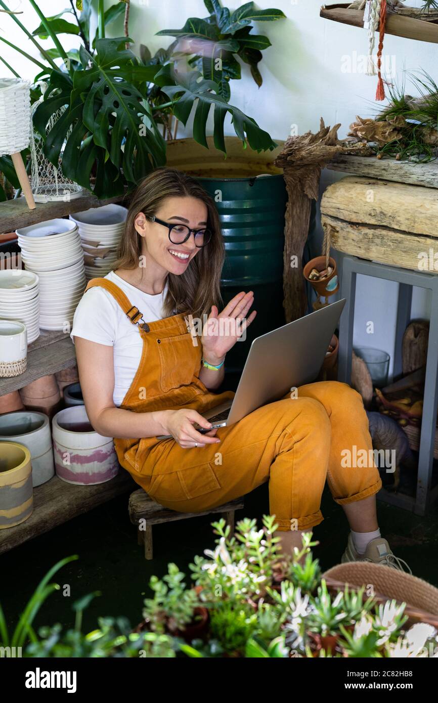 Adorable femme fleuriste en lunettes porter une combinaison orange, assis sur le sol dans un fleuriste, se reposer, utiliser un ordinateur portable après le travail, sourire et parler sur des couleurs vives Banque D'Images