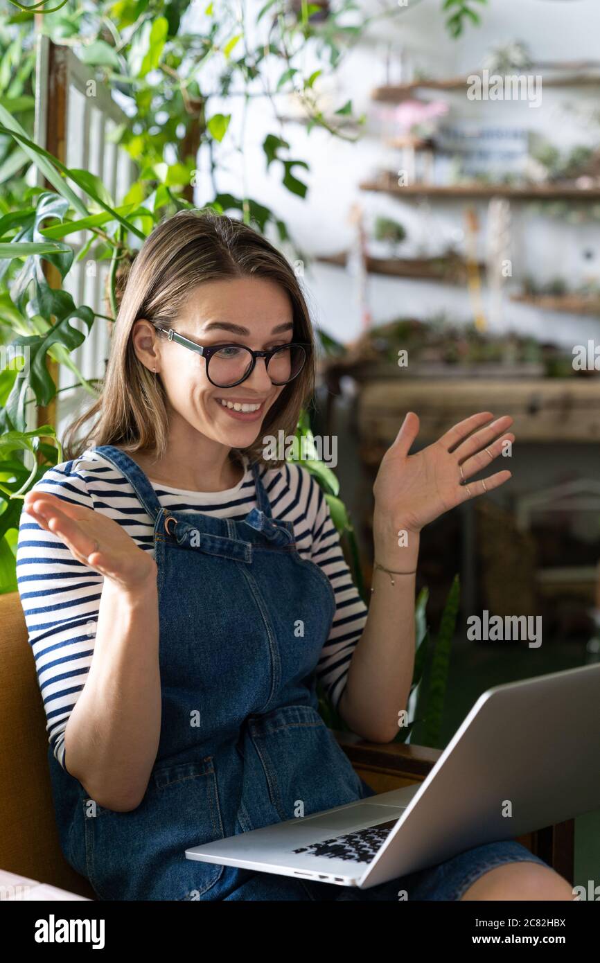 Femme jardinier en lunettes porter bleu jeans combinaison, assis sur une chaise en serre, se reposer, en utilisant un ordinateur portable après le travail, sourire et parler sur la vidéo ca Banque D'Images