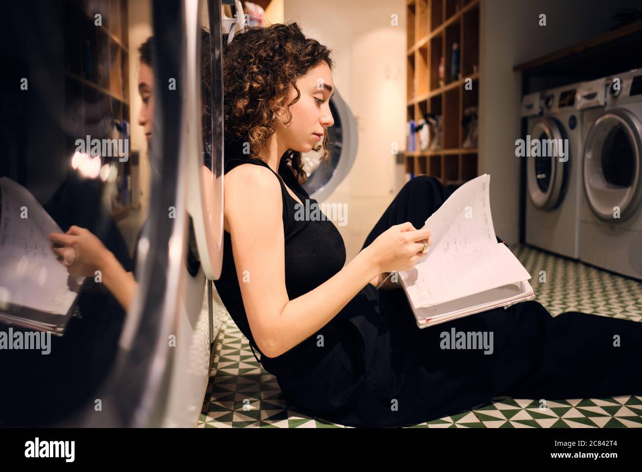 Vue latérale de la fille de brunette lecture de livres en attente de lavage au sol dans la blanchisserie moderne en libre-service Banque D'Images