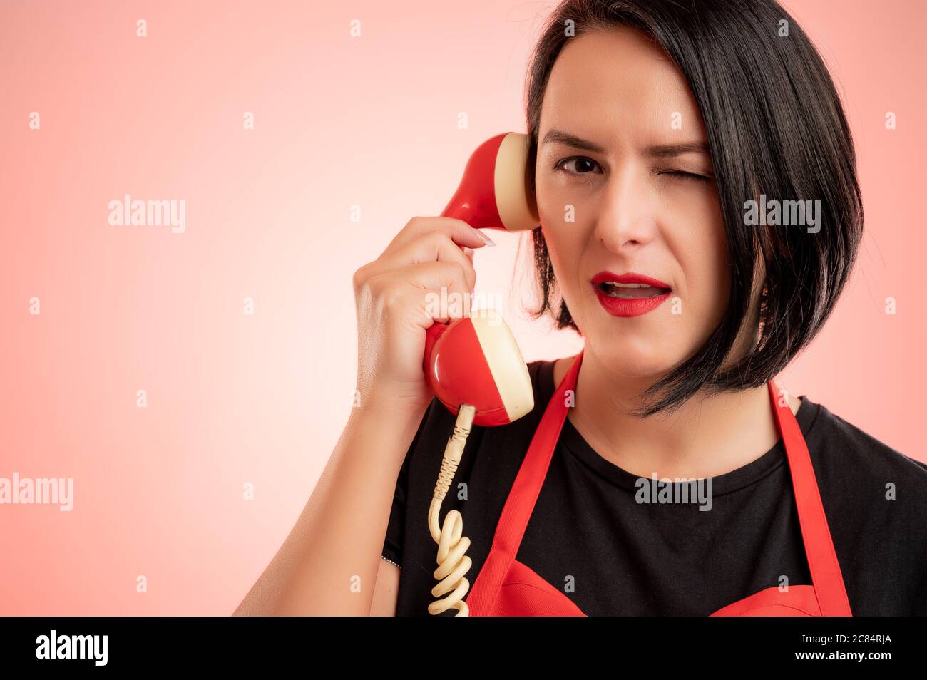 Portrait en gros plan d'une femme de supermarché employée dans un tablier rouge et un t-shirt noir parlant à un ancien récepteur, isolé sur un fond rouge Banque D'Images
