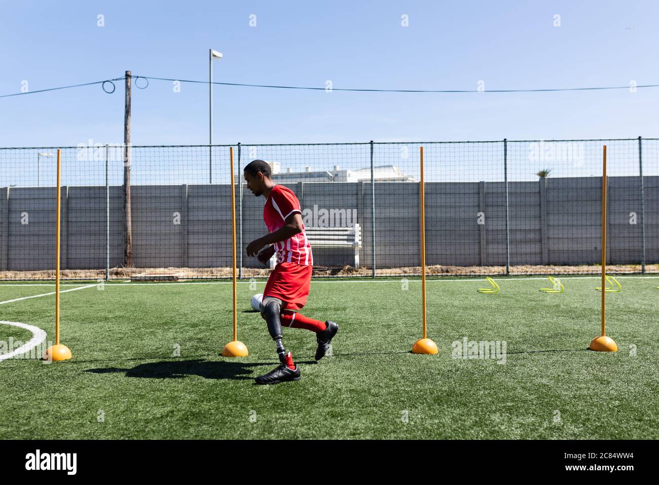 Joueur de football masculin de course mixte avec jambe prothétique portant un entraînement de bande d'équipe sur un terrain de sport au soleil, s'échauffe courir entre les pôles. Banque D'Images