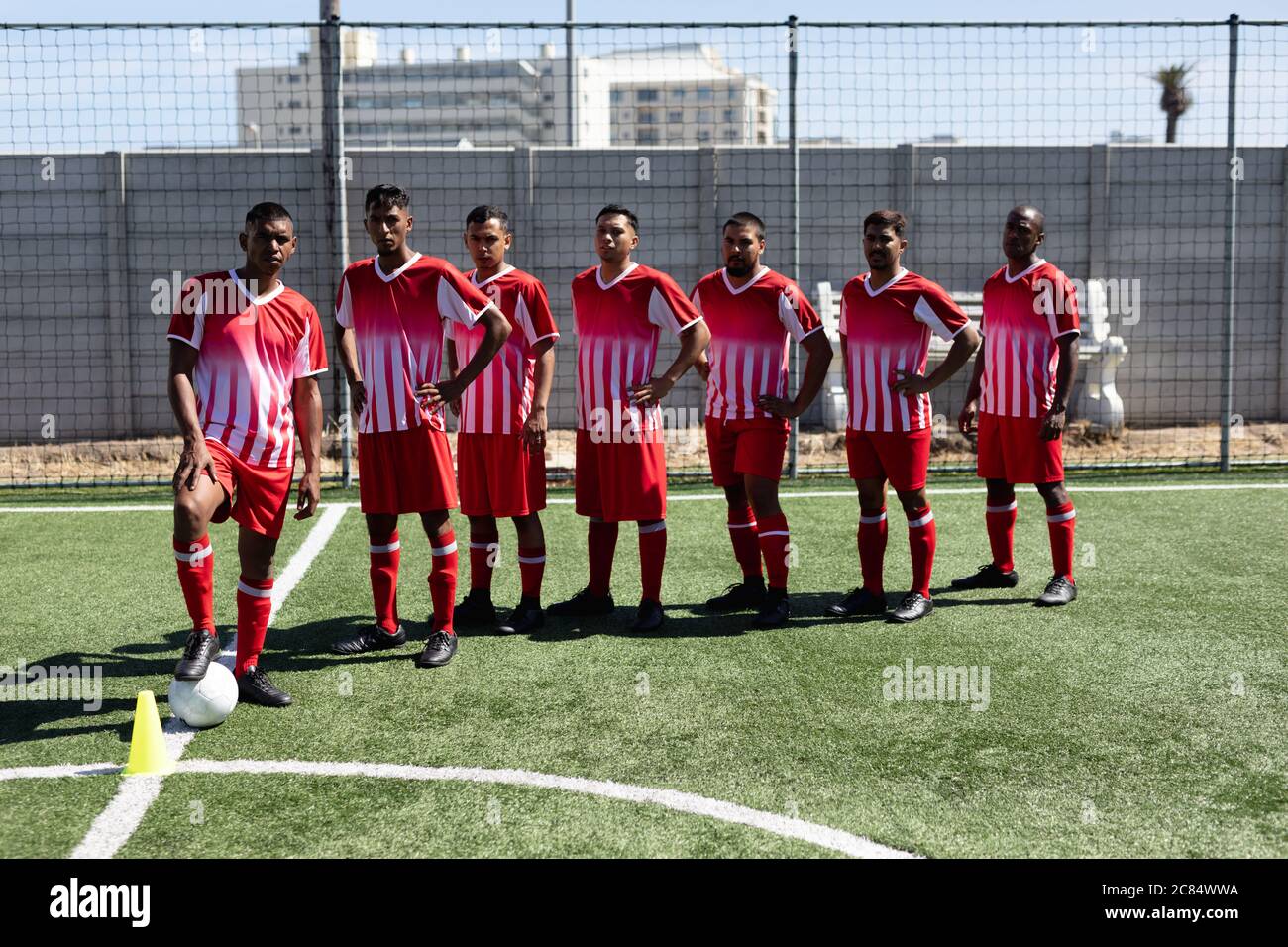 Équipe multi-ethnique de joueurs de football masculins portant un entraînement de bande d'équipe sur un terrain de sport au soleil, posant à la caméra avec une balle. Banque D'Images