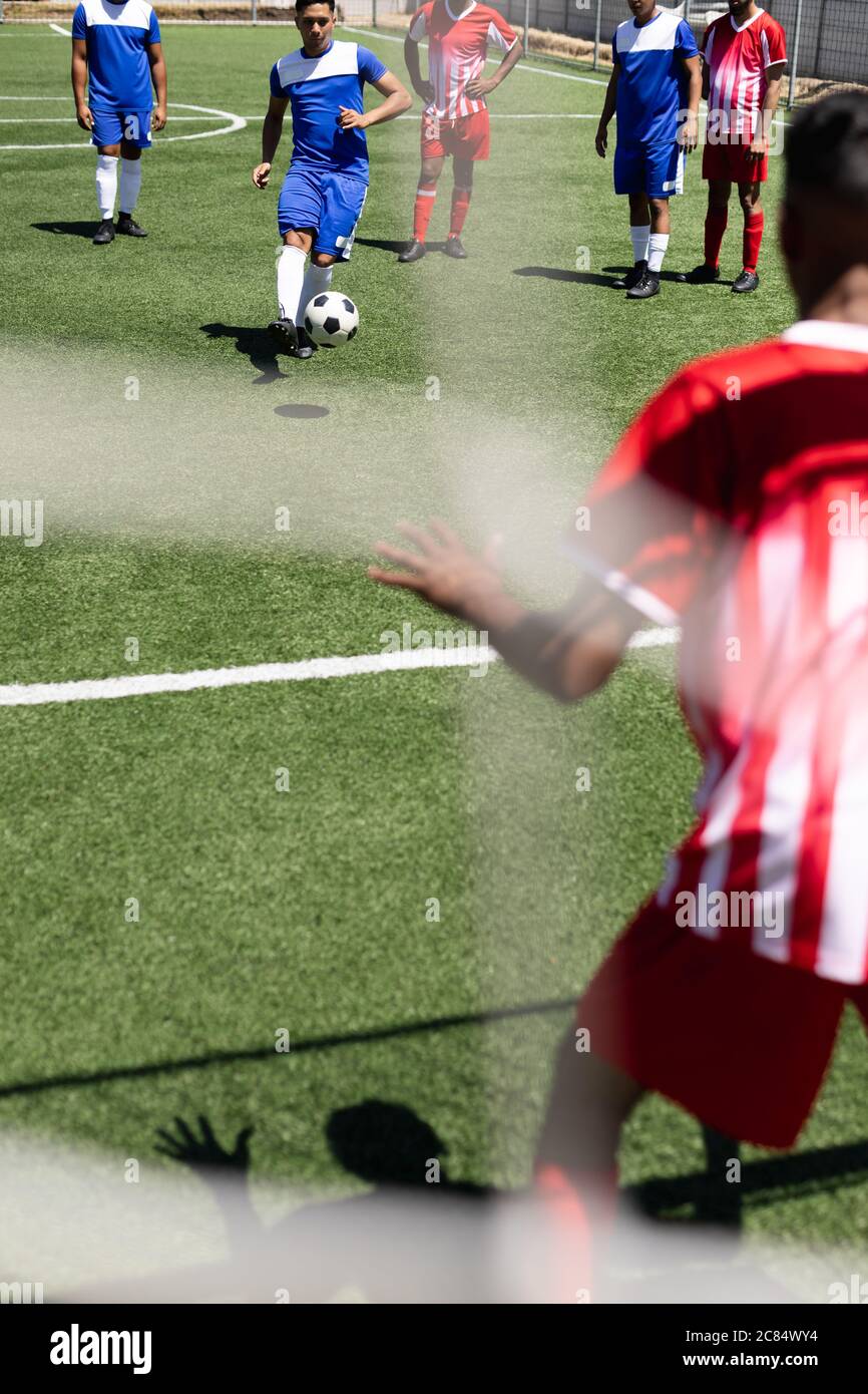 Deux équipes multiethniques de joueurs de football masculins portant une bande d'équipe jouant un match sur un terrain de sport au soleil, un joueur qui donne un coup de pied au gardien de but Banque D'Images