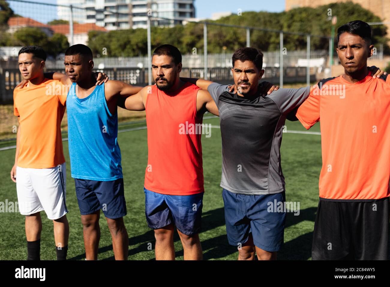 Groupe multiethnique de hommes cinq joueurs de football de côté portant des vêtements de sport entraînement sur un terrain de sport au soleil, debout dans une rangée embrassant befo Banque D'Images