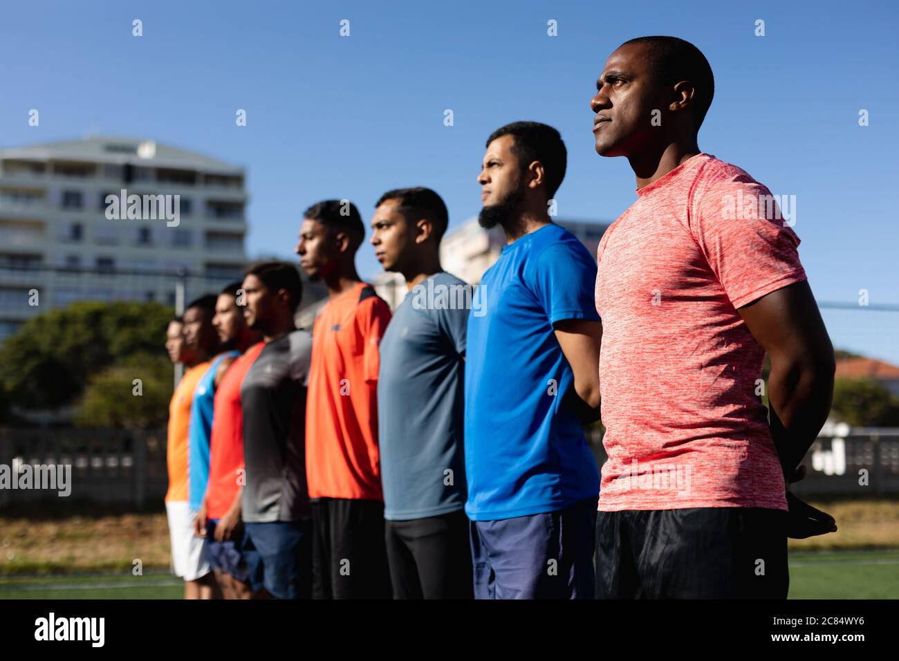 Groupe multiethnique de cinq hommes joueurs de football de côté portant des vêtements de sport entraînement sur un terrain de sport au soleil, debout dans une rangée avant un match. Banque D'Images