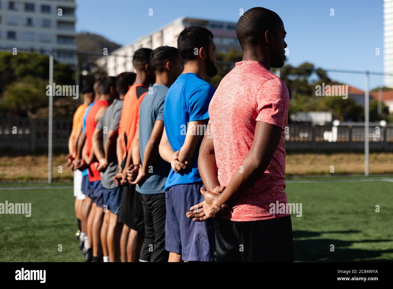 Groupe multiethnique de cinq hommes joueurs de football de côté portant des vêtements de sport entraînement sur un terrain de sport au soleil, debout dans une rangée avant un match. Banque D'Images