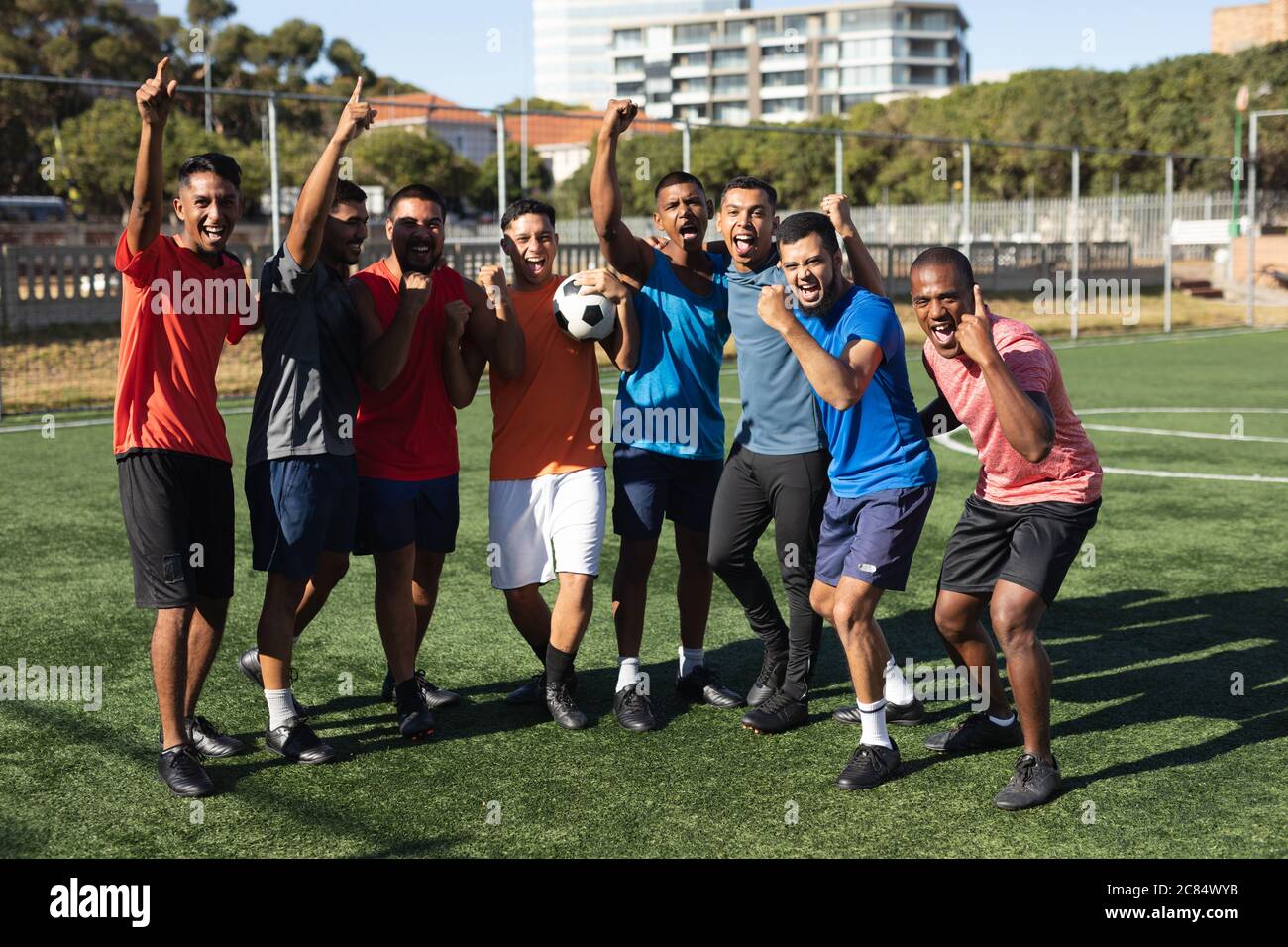 Groupe multiethnique de cinq hommes joueurs de football de côté portant des vêtements de sport entraînement sur un terrain de sport au soleil, célébrant la victoire tenue d'un ba Banque D'Images
