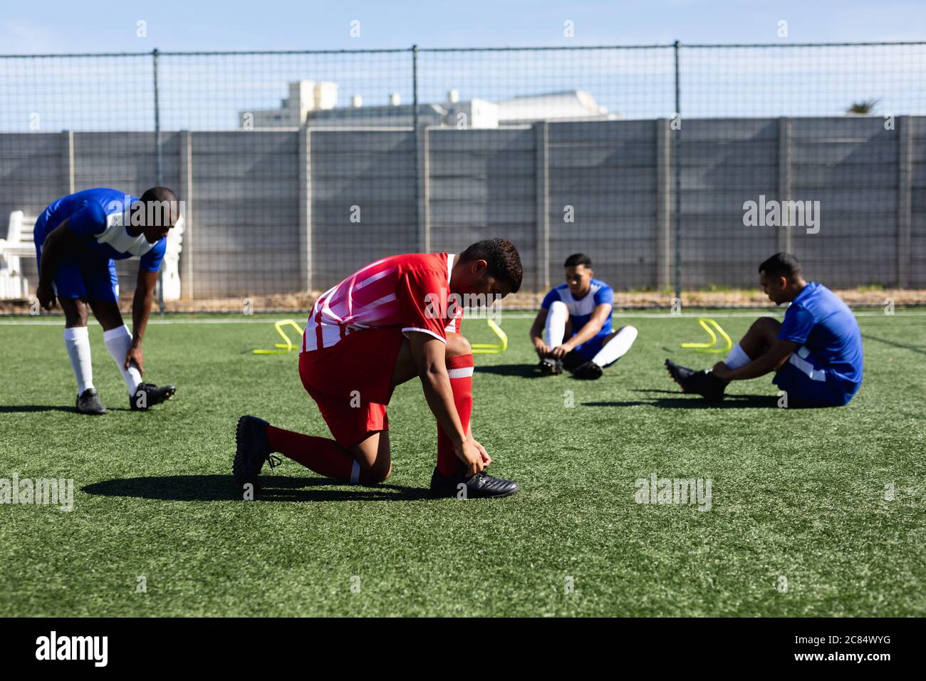 Multiethnique masculin cinq joueurs de football de côté portant un entraînement de bande d'équipe sur un terrain de sport au soleil, en se réchauffant de leurs lacets. Banque D'Images
