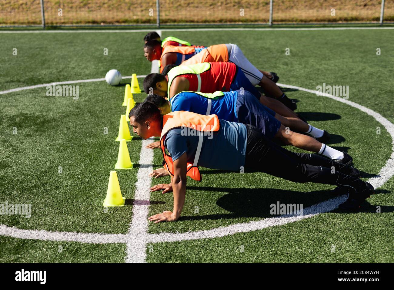 Groupe multiethnique de hommes cinq joueurs de football de côté portant des vêtements de sport et des gilets de sport s'entraîner sur un terrain de sport au soleil, en se réchauffant en faisant pousser Banque D'Images
