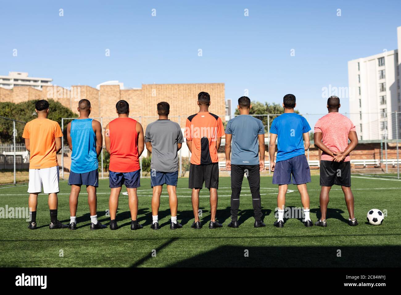 Vue arrière d'un groupe multiethnique de cinq hommes joueurs de football de côté portant des vêtements de sport entraînement sur un terrain de sport au soleil, debout dans une rangée b Banque D'Images