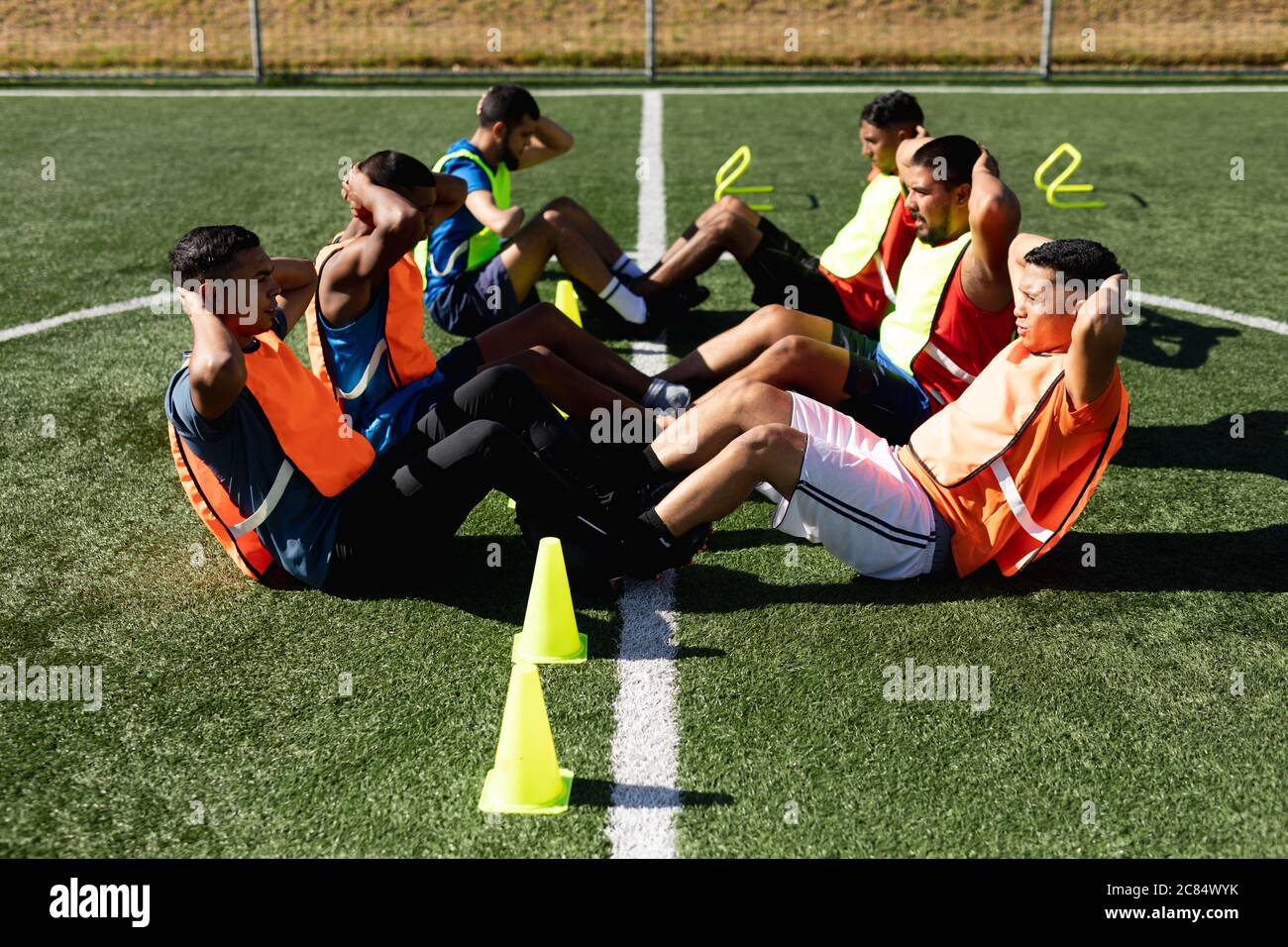 Groupe multiethnique de hommes cinq joueurs de football de côté portant des vêtements de sport et des gilets de sport s'entraîner sur un terrain de sport au soleil, en se réchauffant en faisant sit u Banque D'Images