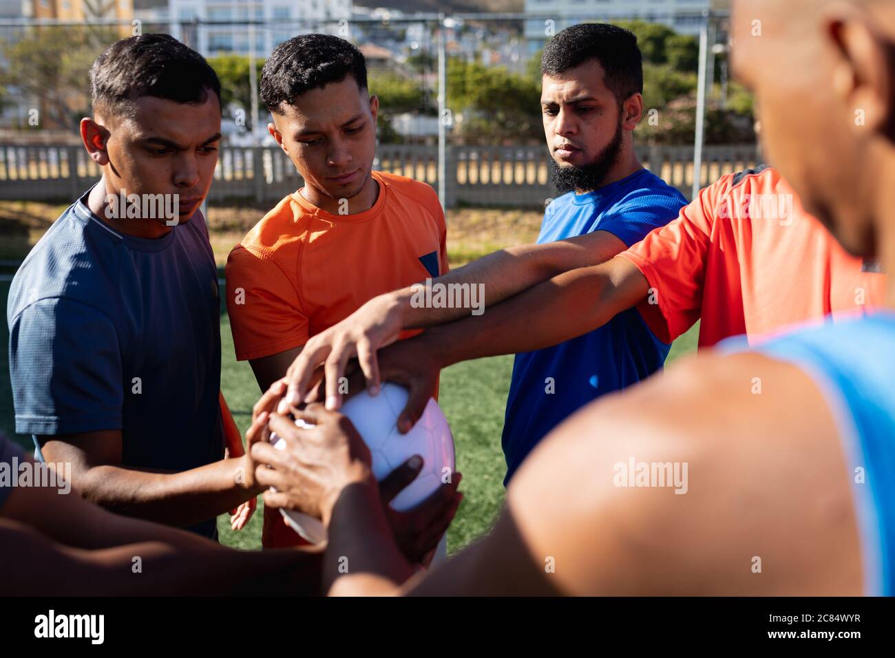 Groupe multiethnique de cinq hommes joueurs de football de côté portant des vêtements de sport entraînement sur un terrain de sport au soleil, debout la main s'empilant sur un ballon Banque D'Images