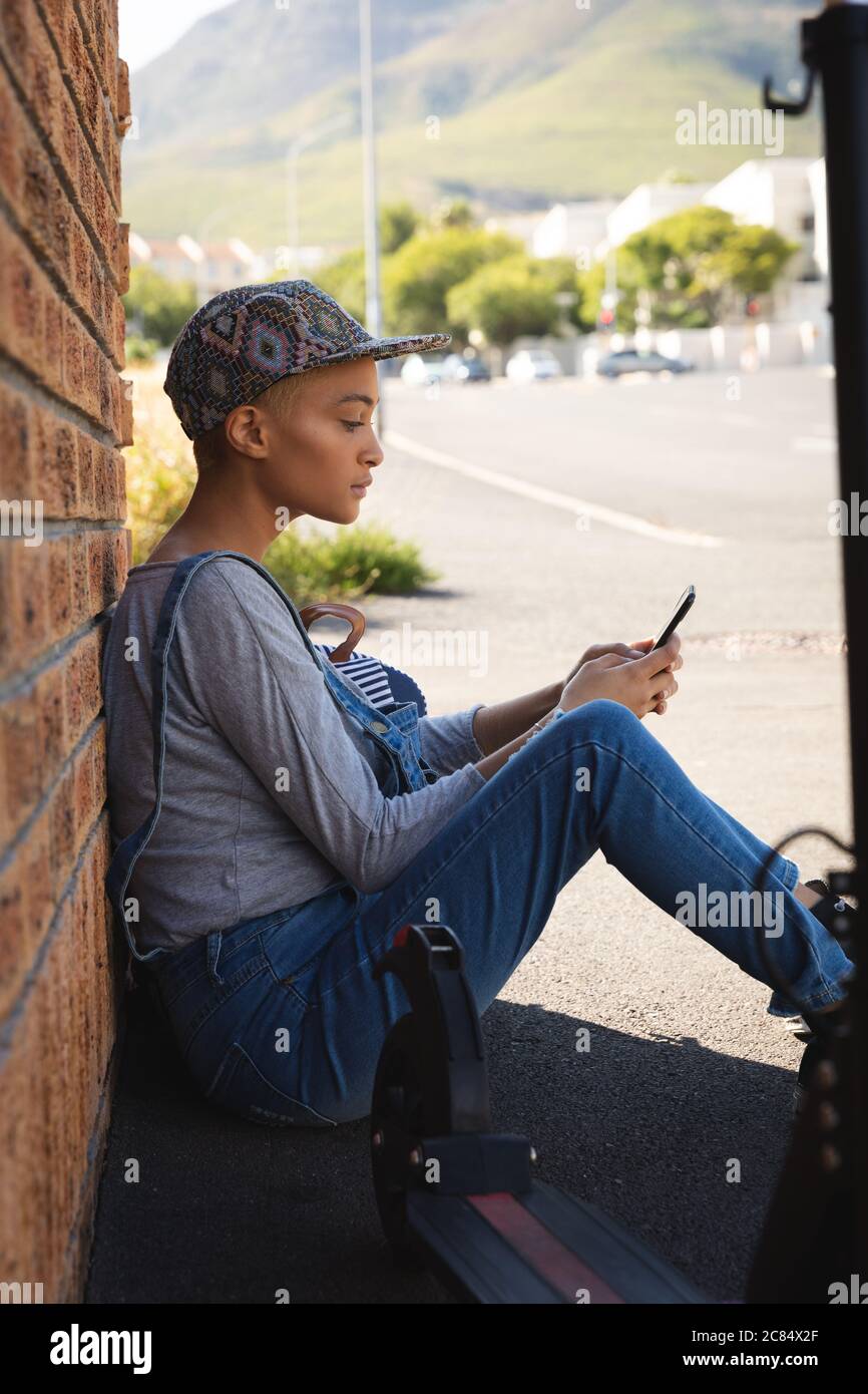 Femme alternative de course mixte avec cheveux courts dehors et autour dans  la ville un jour ensoleillé, portant des salopettes en denim et une  casquette, assis contre le mur avec des s