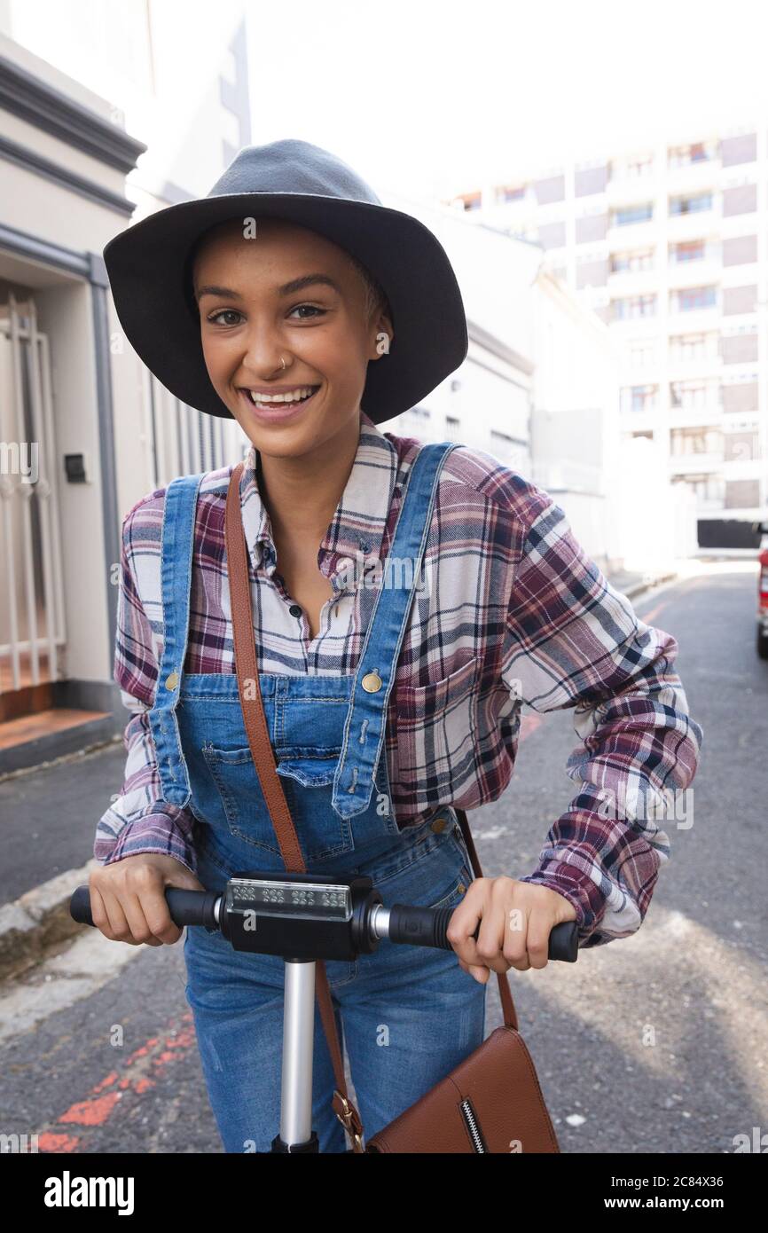 Une alternative de course mixte femme avec des cheveux courts dehors et  autour dans la ville sur un jour ensoleillé, portant un chapeau et  salopette, en utilisant un scooter et souriant à
