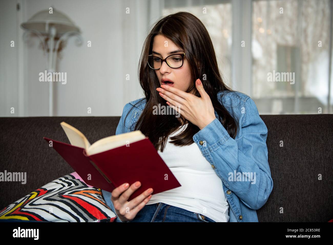 Une jeune femme choquée avec un livre assis sur un canapé à la maison a choqué une jeune femme avec un livre assis sur un canapé à la maison Banque D'Images