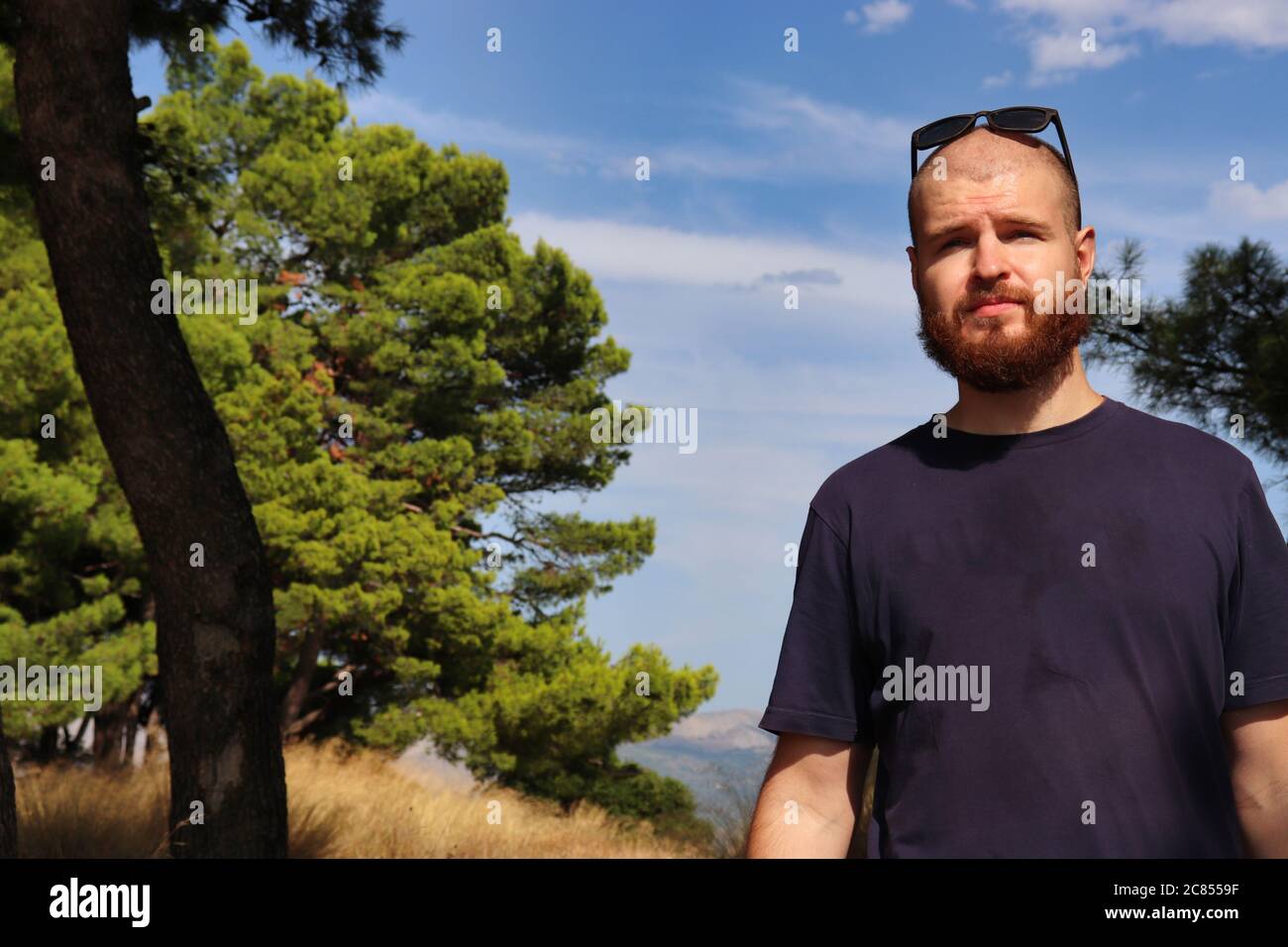 Jeune adulte à tête blanche barbu avec des lunettes de soleil sur sa tête debout à l'extérieur dans la nature avec une expression inquiète regardant dans la distance Banque D'Images