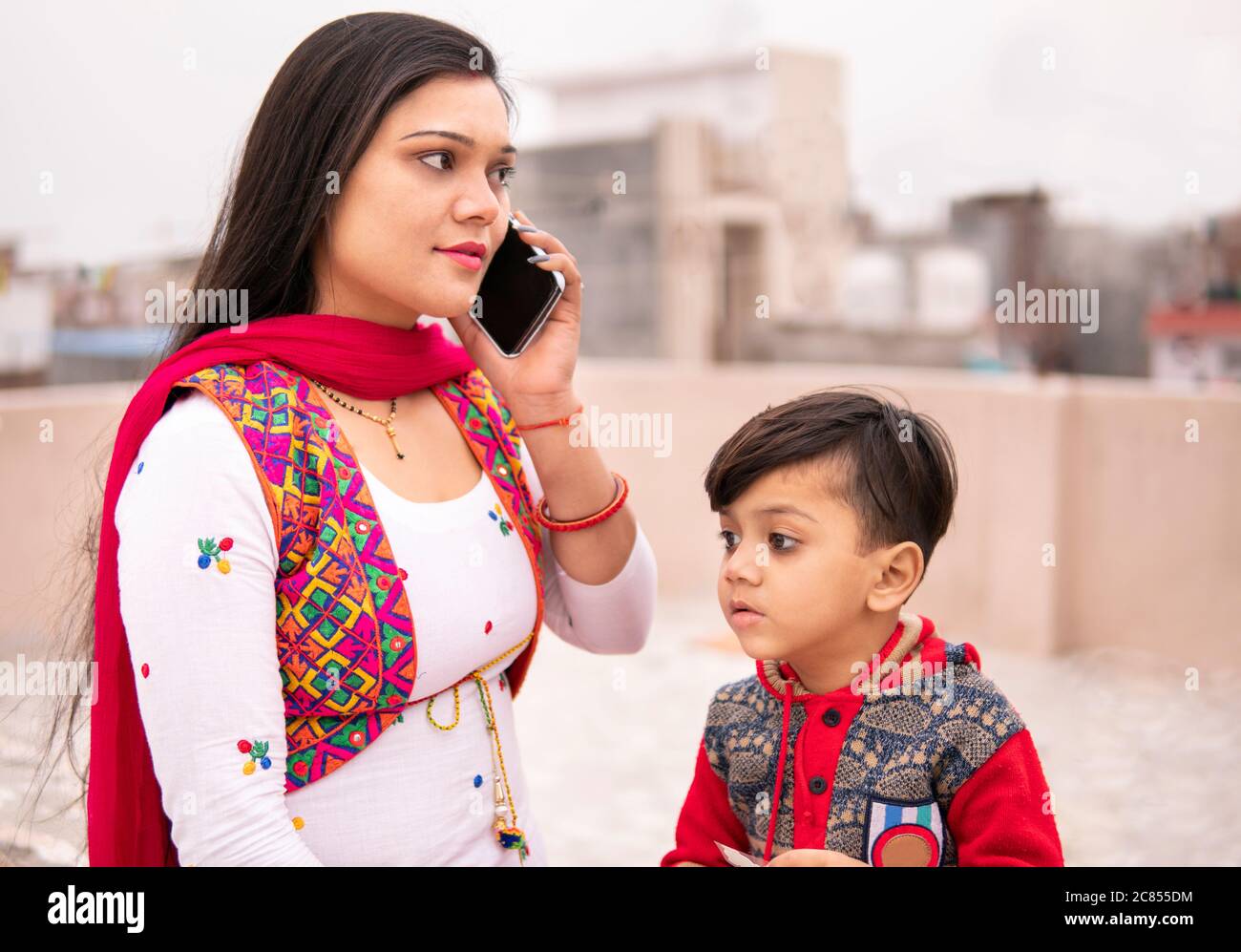 Belle et heureuse jeune mère indienne avec son fils sur le toit et parlant sur un smartphone à l'heure de la journée. Elle porte la robe traditionnelle salwar Kameez. Banque D'Images