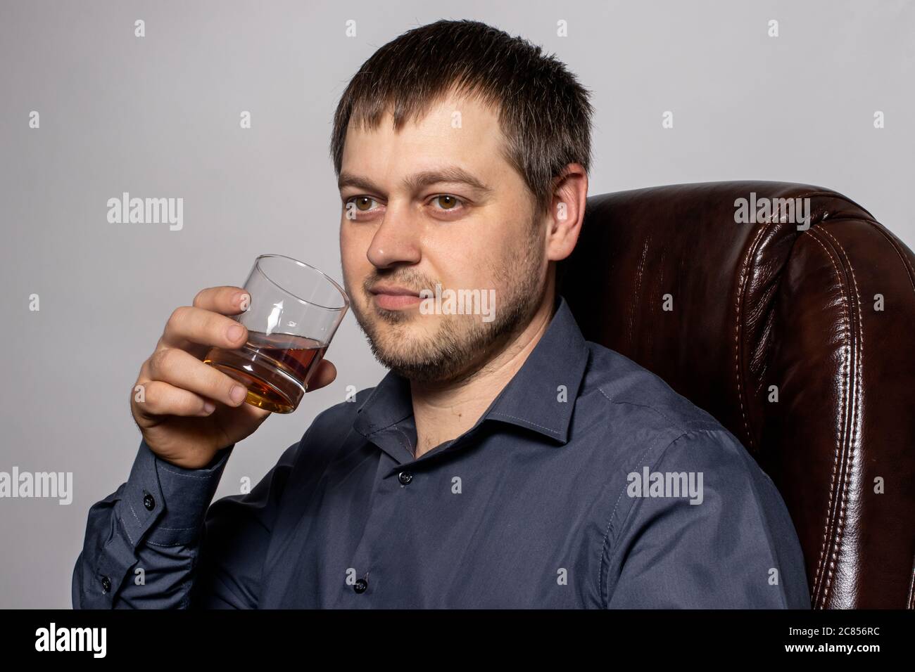 Un beau jeune homme de 30-35 ans dans une chemise est assis sur un fauteuil en cuir marron et tient un verre de whisky dans la main. Banque D'Images