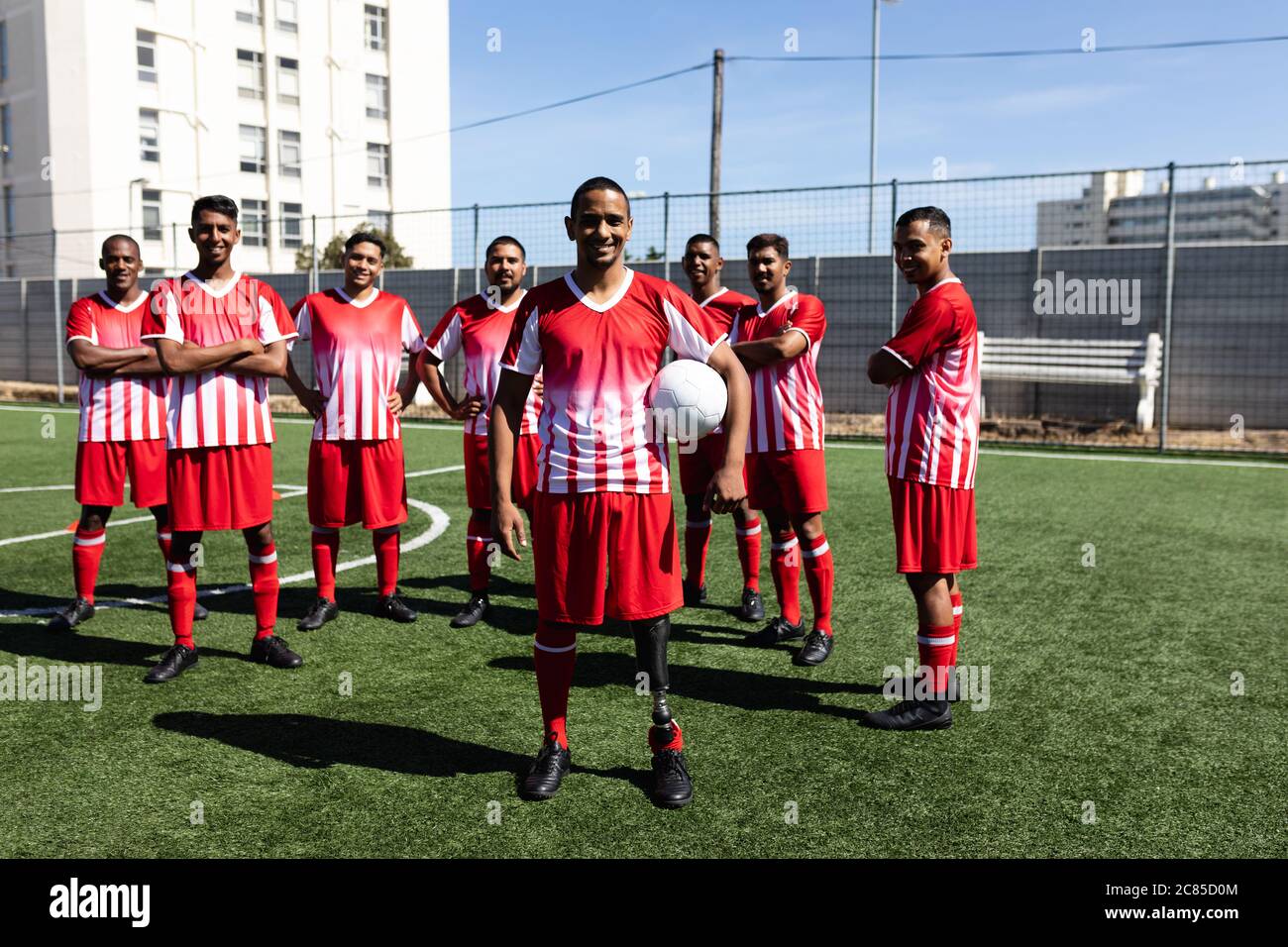 Portrait d'une équipe multiethnique de cinq hommes joueurs de football de côté portant un entraînement de bande d'équipe sur un terrain de sport au soleil, debout tenant une balle Banque D'Images