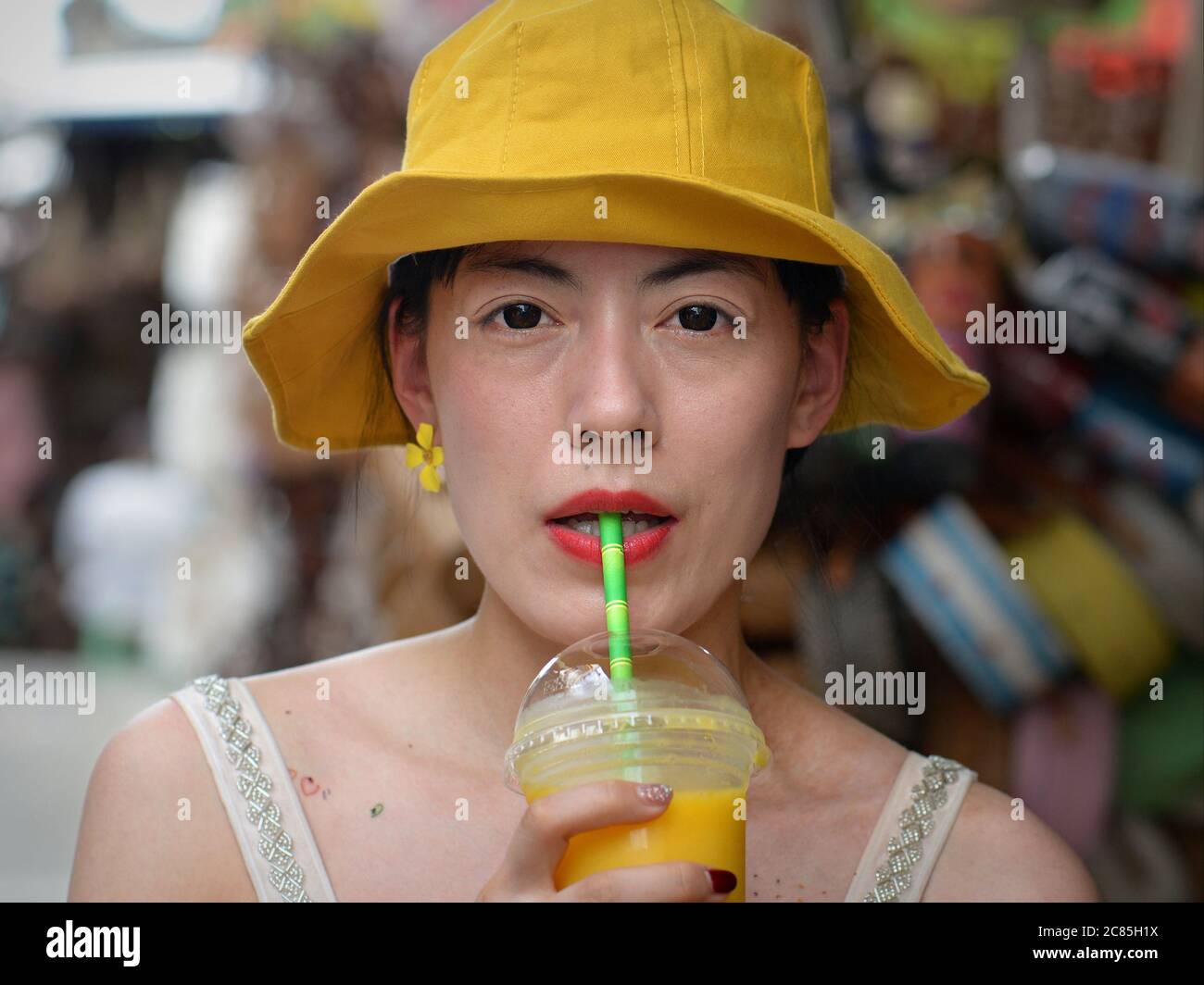Jeune femme asiatique avec chapeau de disquettes jaune porte des lentilles de contact foncées et boit du jus de fruits jaunes avec une paille d'une tasse en plastique. Banque D'Images