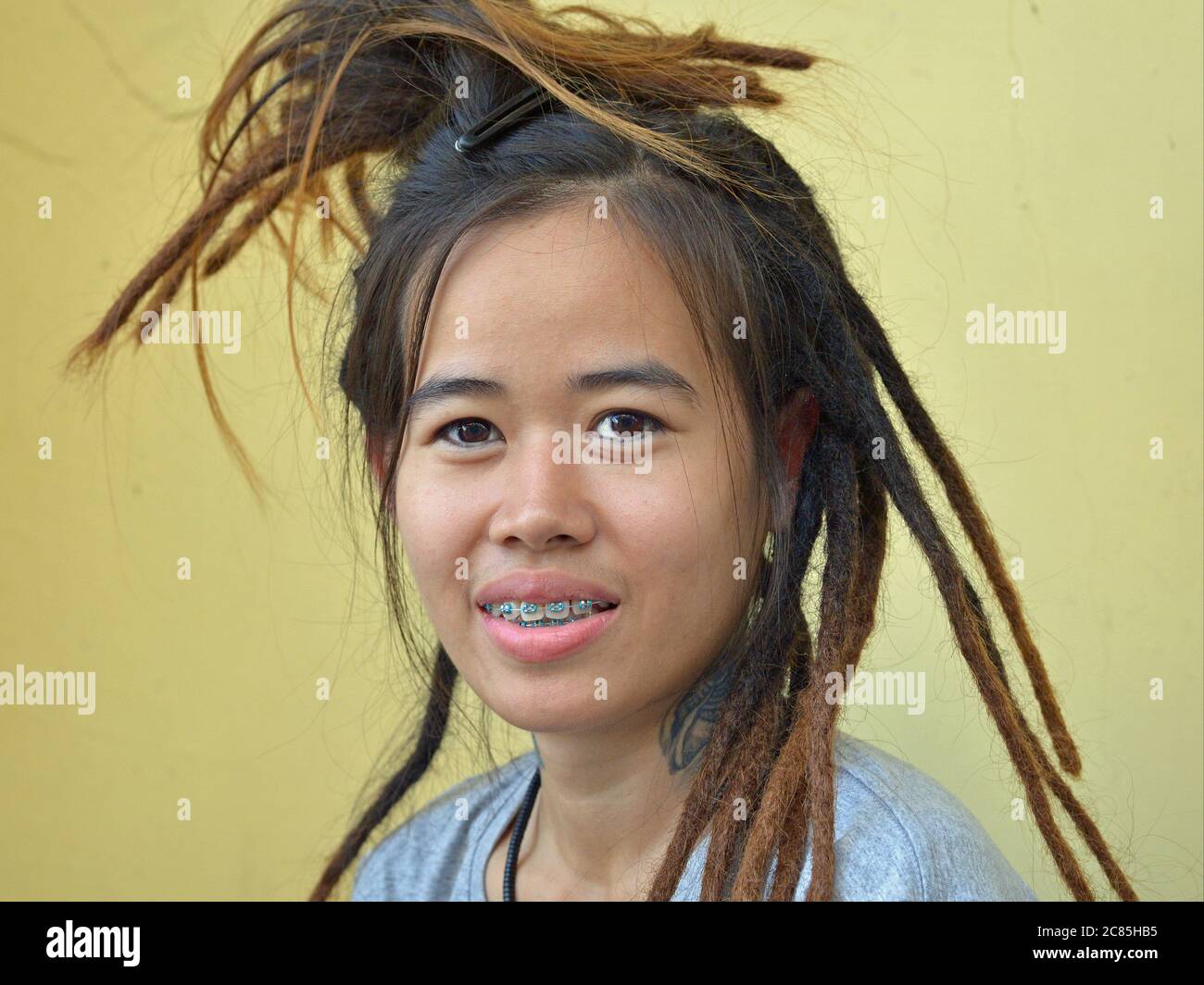 Jeune femme thaïlandaise avec des bretelles dentaires bleues et longue, seulement la moitié finie Rastafarian relit sourire pour la caméra. Banque D'Images
