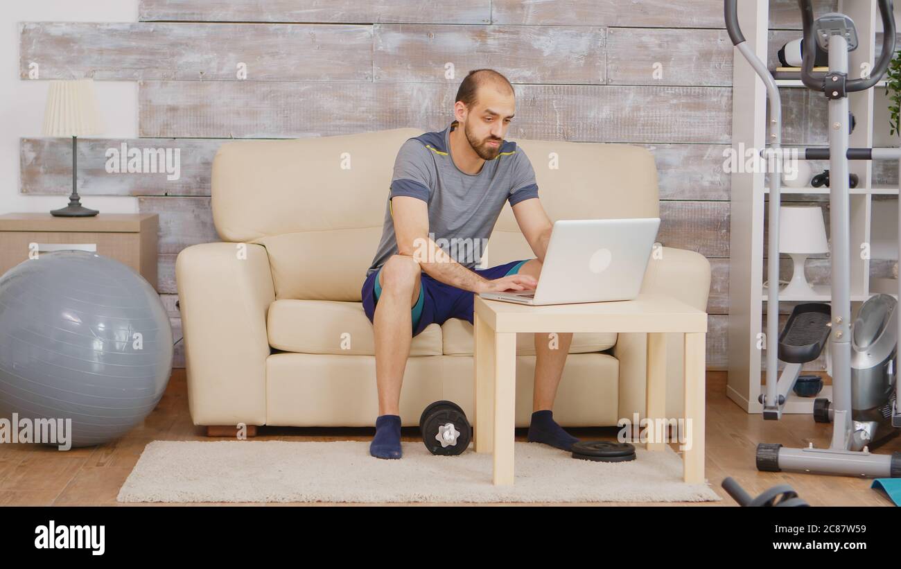 Guy dans les vêtements de sport recherchant entraînement biceps avec haltères sur ordinateur portable assis sur le canapé. Banque D'Images