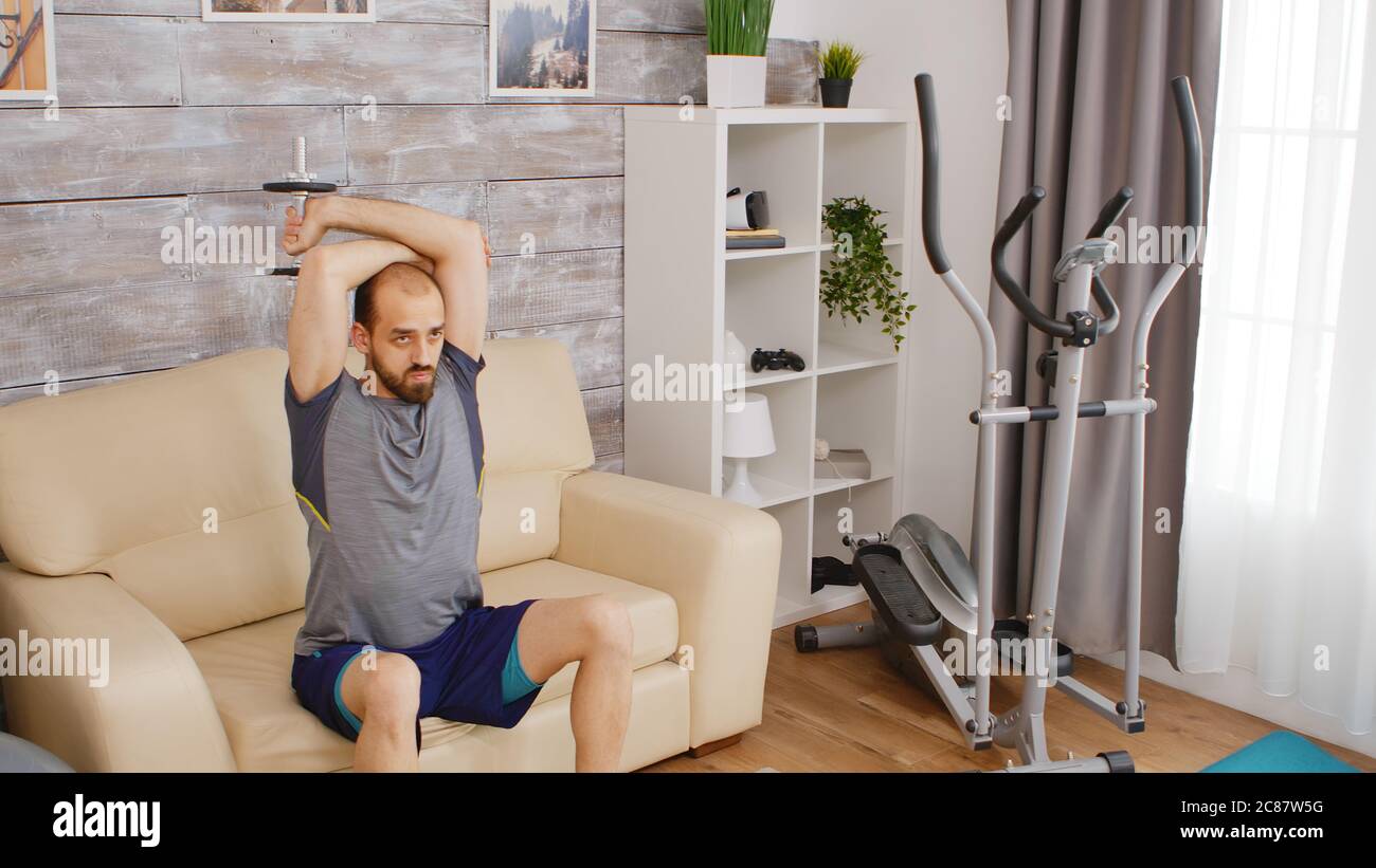 Homme sportif entraînement triceps muscle avec haltères assis sur le canapé dans le salon. Banque D'Images