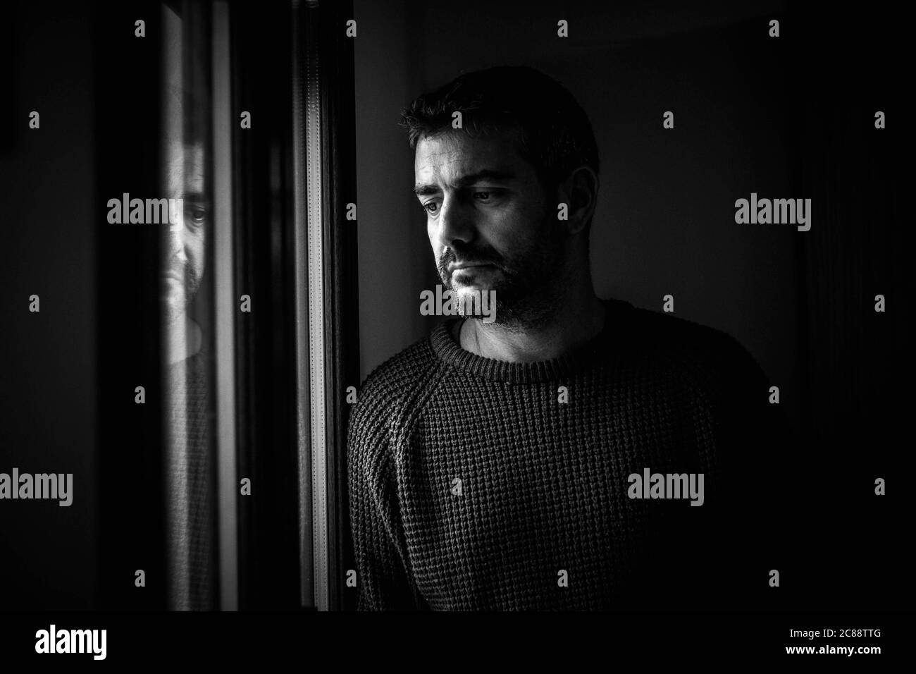 Prise de vue en niveaux de gris d'un homme d'âge moyen du caucase debout près du fenêtre avec un regard triste et bien pensé Banque D'Images
