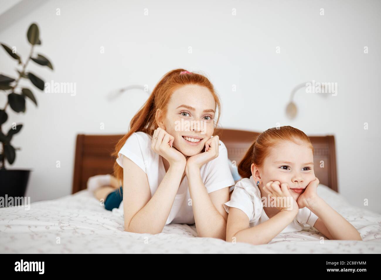 Jeune mère aux cheveux rouges à la maison, vêtir avec sa petite fille de quatre ans regardant l'appareil photo tout en étant allongé sur un lit dans une chambre blanche. Profitez de votre temps libre Banque D'Images