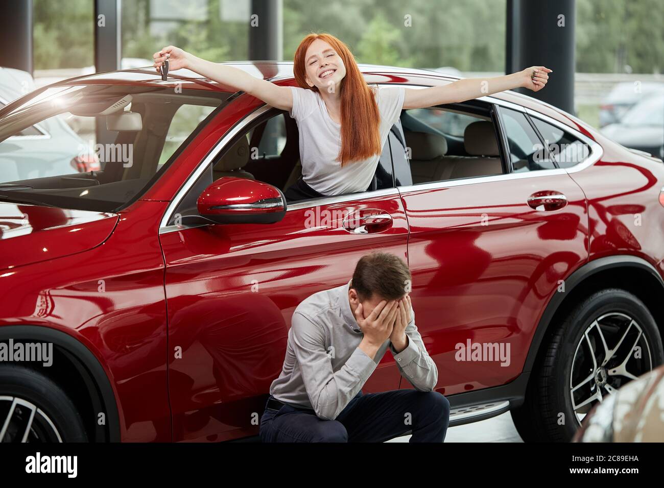 L'achat d'une nouvelle voiture provoque des émotions divergentes. Une femme heureuse aux cheveux rouges est surjoyeuse et se tient hors de la voiture avec les mains qui se répandent et son mari anxieux Banque D'Images