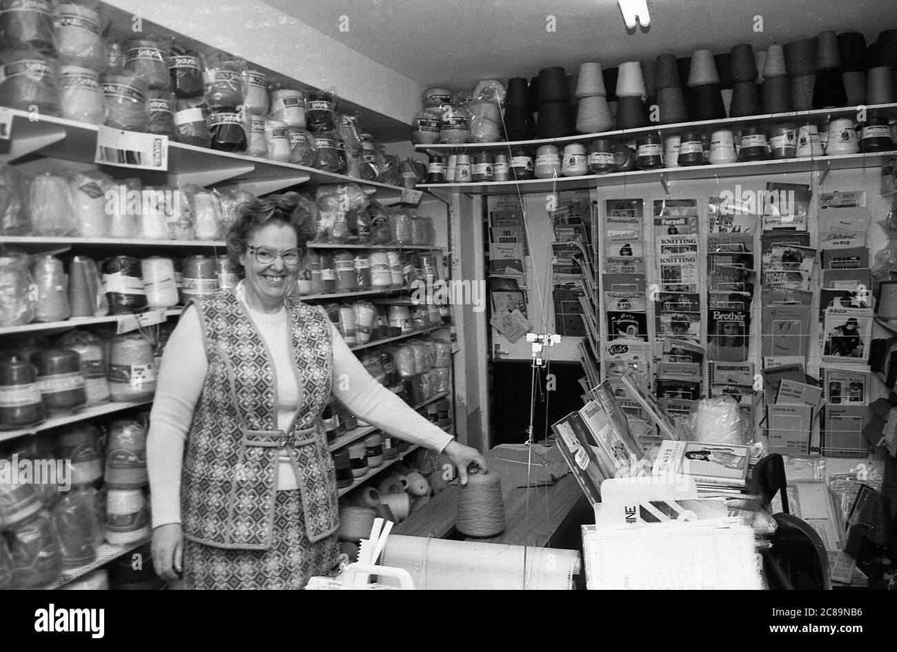 Années 1980, propriétaire historique, femme d'un magasin vendant de la laine debout dans son magasin, montrant la grande gamme de différents laines disponibles et tricots magazines Angleterre, Royaume-Uni. Banque D'Images