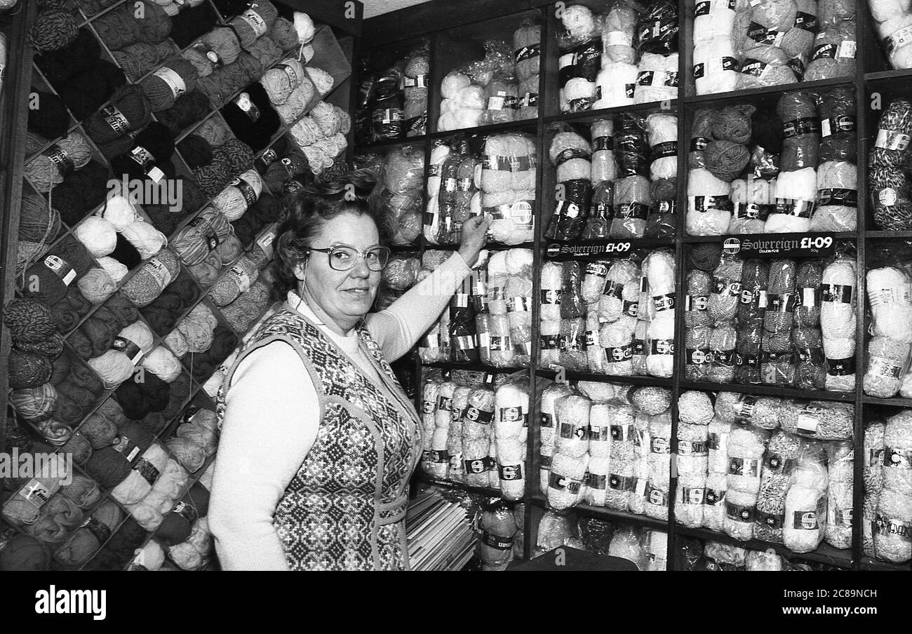 Années 1980, propriétaire historique, femme d'un magasin vendant de la laine debout par des shevles pleins de stock, Angleterre, Royaume-Uni. Banque D'Images