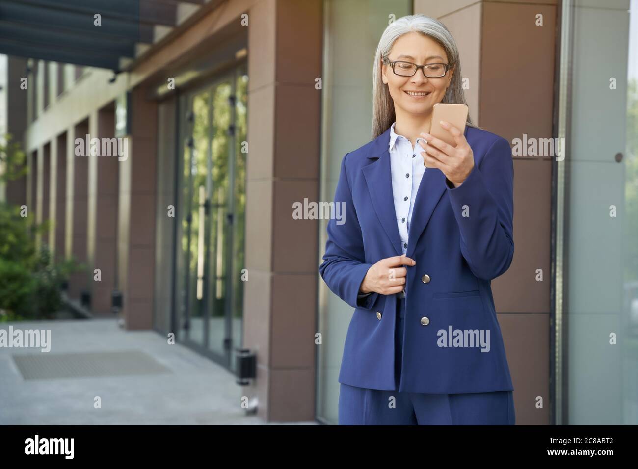 Portrait d'une femme d'affaires mûre et attrayante portant un costume classique et des lunettes de vue à l'aide d'un téléphone portable et souriant tout en se tenant debout contre le bâtiment de bureau à l'extérieur. Les gens d'affaires Banque D'Images