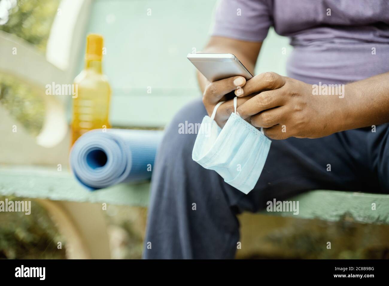 Homme utilisant le mobile avec le masque à la main au parc pendant le yoga du matin ou l'entraînement avec mat - concept de coronavirus ou Covid-19 conseils, protection ou à Banque D'Images