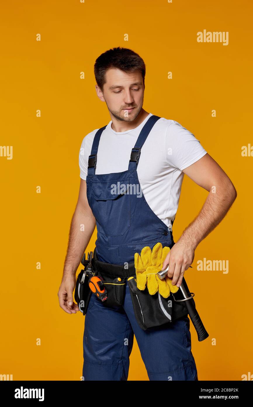 Jeune réparateur caucasien en casque dur jaune et en marteau de maintien uniforme. Handyman portant une ceinture à outils isolée sur fond orange studio Banque D'Images