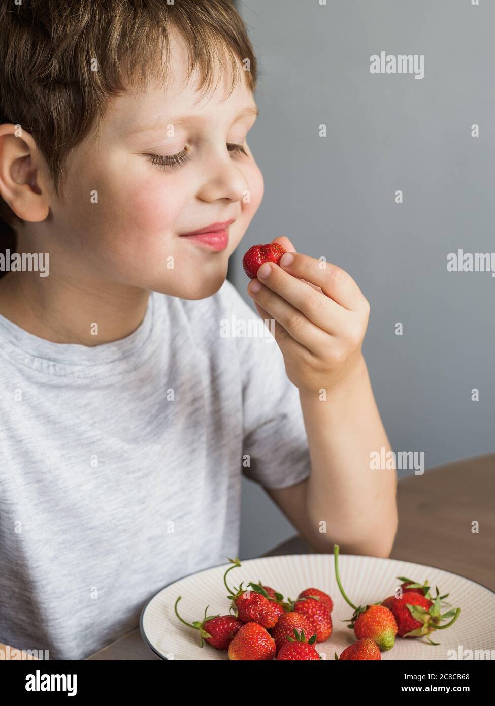 Un garçon de quatre ans mange des fraises fraîches avec de la relish. Un enfant souriant mange des fraises bio à la table de la cuisine. Verticale Banque D'Images