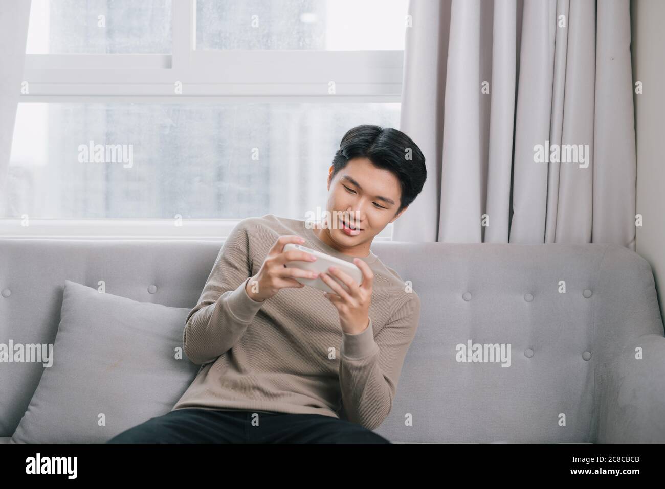 Le jeune homme asiatique joue sérieusement sur son smartphone sur le canapé. Banque D'Images
