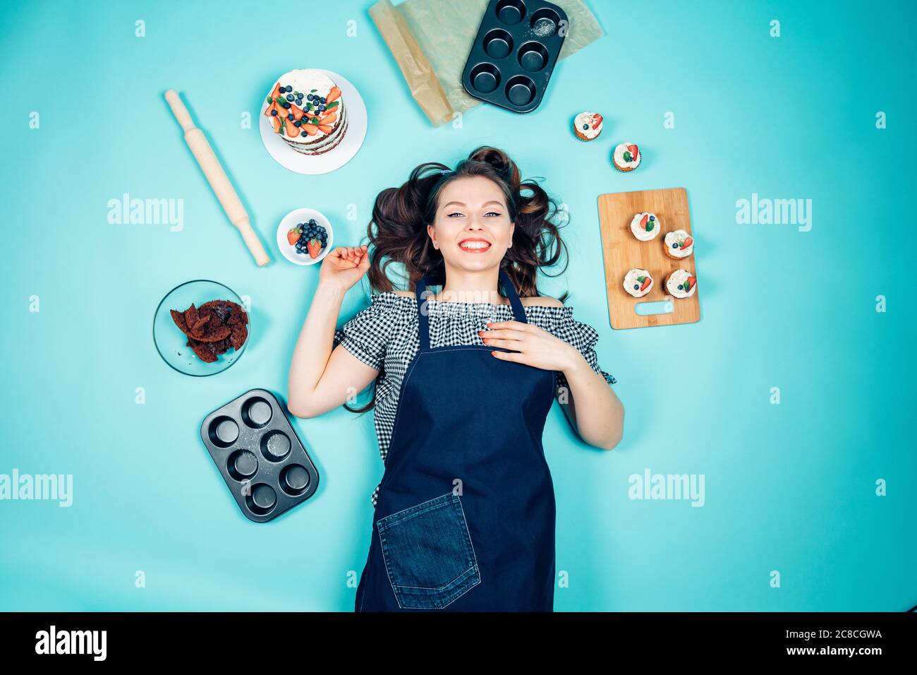 Souriant, le joli chef pâtissier avec des gâteaux et des ingrédients pour cuisiner des gâteaux couchés sur le sol, regardant un appareil photo Banque D'Images