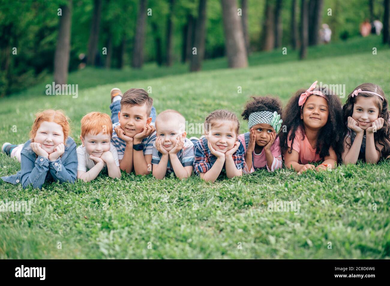 Groupe d'enfants positifs en âge d'aller à l'école primaire, allongé sur de l'herbe verte dans le parc. Le concept de l'amitié, de l'enfance et de la communication interculturelle. Banque D'Images